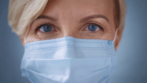 κοντινό πλάνο μεσήλικης γυναίκας με μπλε μάτια σε ιατρική μάσκα κοιτάζοντας την κάμερα - Πλάνα, βίντεο