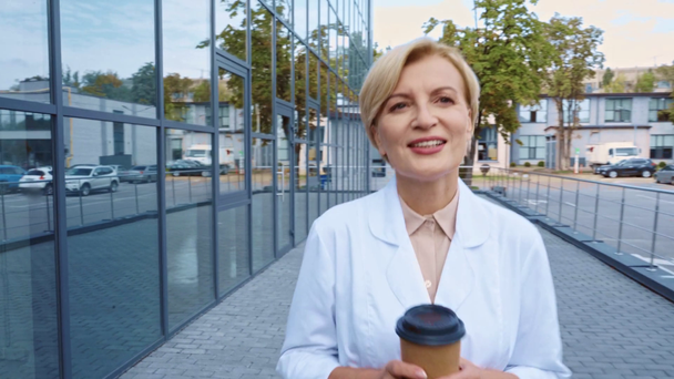 tevreden arts van middelbare leeftijd die papieren beker met koffie vasthoudt en buiten loopt - Video