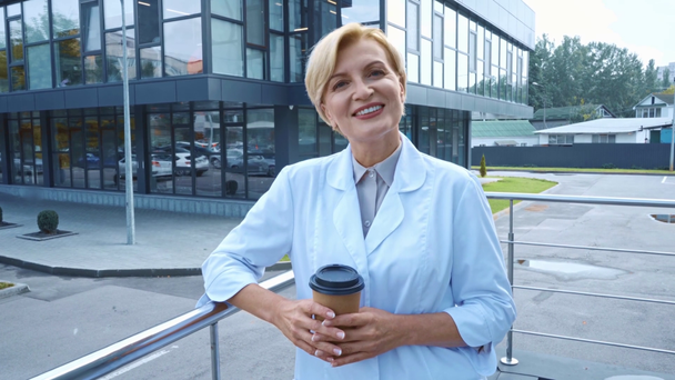 pleased middle aged doctor holding paper cup with coffee outside - Footage, Video