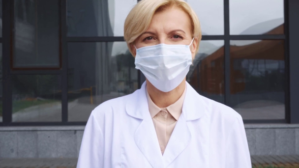középkorú orvos fehér köpenyben és orvosi maszkban kint áll - Felvétel, videó
