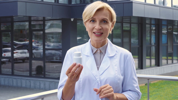 middle aged doctor in white coat holding bottle with pills and talking outside - Filmagem, Vídeo