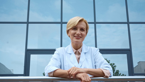 szczęśliwy lekarz w średnim wieku w białym płaszczu stojący w pobliżu budynku ze szklaną fasadą - Materiał filmowy, wideo