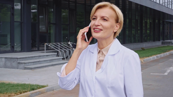 μεσήλικας γιατρός με λευκό παλτό περπατώντας έξω και μιλώντας στο smartphone - Πλάνα, βίντεο