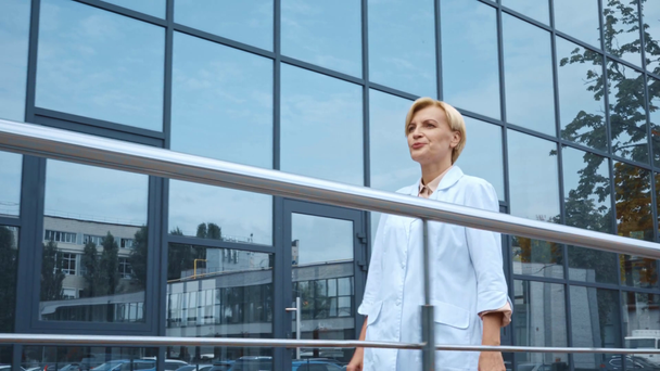 gelukkige dokter van middelbare leeftijd in witte jas wandelen in de buurt van gebouw met glazen gevel - Video