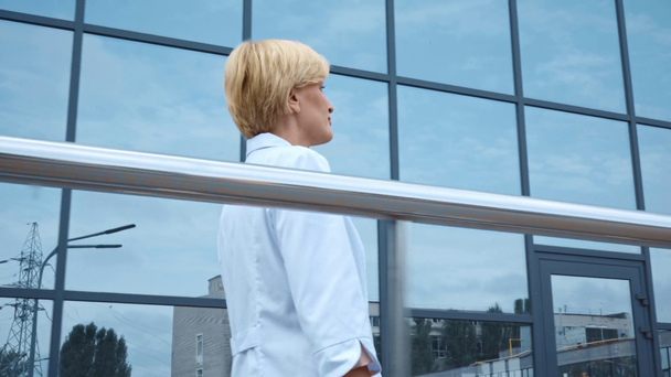 médico de meia idade loira em casaco branco andando perto do edifício com fachada de vidro - Filmagem, Vídeo