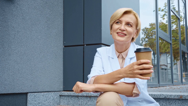 sonriente médico de mediana edad en abrigo blanco sosteniendo taza de papel afuera  - Metraje, vídeo