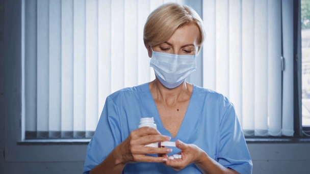 μεσήλικας γιατρός σε ιατρική μάσκα άνοιγμα μπουκάλι και δείχνει χάπια  - Πλάνα, βίντεο