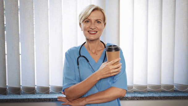 alegre médico de mediana edad sosteniendo taza de papel con café en la clínica  - Metraje, vídeo