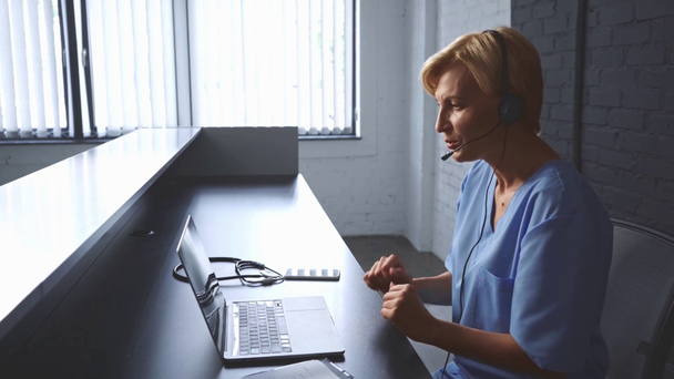 lékař středního věku ve sluchátkách poradenství on-line v blízkosti notebooku na klinice - Záběry, video