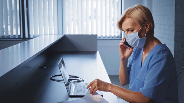 middle aged doctor in medical mask typing on laptop and talking on smartphone - Footage, Video