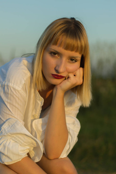 A vertical shot of a beautiful caucasian woman wearing a white shirt, sitting on a stool outdoors - Fotó, kép