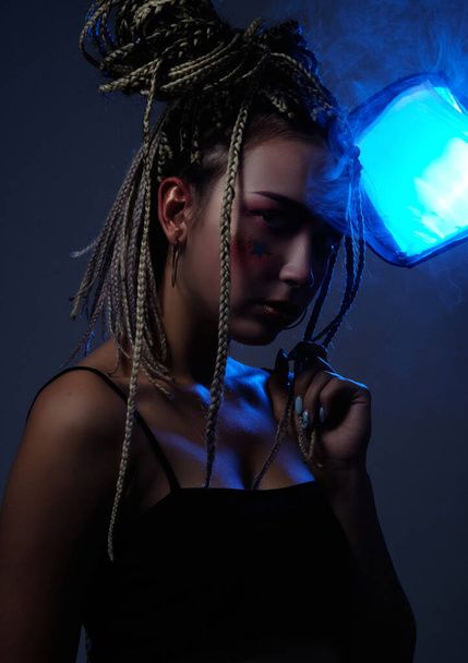 girl with dreadlocks in a black top with creative makeup posing in the studio with blue light - Valokuva, kuva