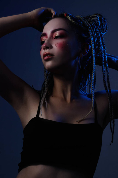 girl with dreadlocks in a black top with creative makeup posing in the studio with blue light - Photo, Image