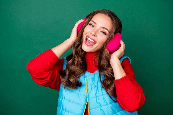 Photo of pretty cute young lady dreamy look open mouth pretend listen music favorite singer sing song wear pink ear warmers blue vest red sweater isolated green color background - Foto, Imagen