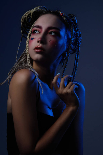 girl with dreadlocks in a black top with creative makeup posing in the studio with blue light - Фото, изображение