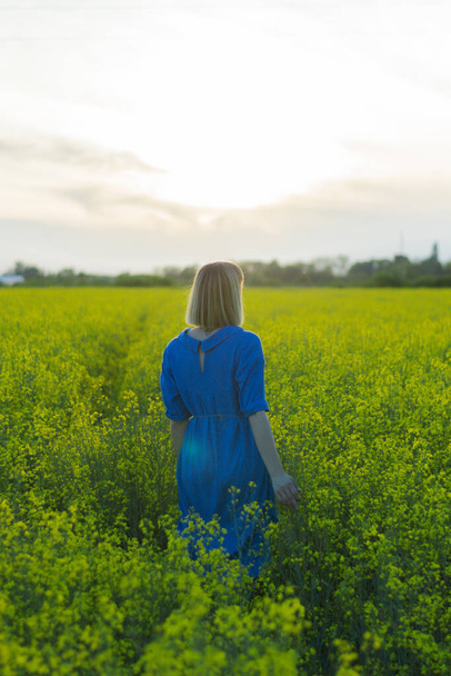 A vertical shot of a beautiful caucasian woman wearing a blue dress, standing in a field full of yellow flowers during daylight - Fotografie, Obrázek