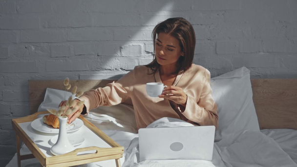 Junge Freiberuflerin trinkt Kaffee und tippt auf Laptop im Bett - Filmmaterial, Video
