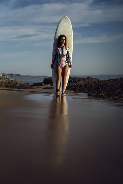 A vertical shot of a young female in a swimsuit holding a surfboard on the beach - Фото, изображение