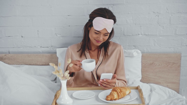 брюнетка женщина пьет кофе и сообщения на смартфоне в постели - Кадры, видео