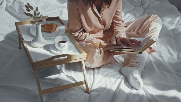 vista recortada de la mujer bebiendo café y leyendo libro en la cama - Imágenes, Vídeo