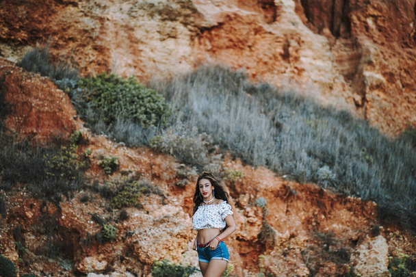 A sexy caucasian woman in short jeans and an open shirt showing a fit belly standing with an immense cliff behind - Fotografie, Obrázek