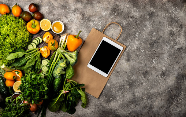 Tablet υπολογιστή και τσάντα χαρτί ψώνια με διαφορετικά βιολογικά λαχανικά και χόρτα. Άνω όψη - Φωτογραφία, εικόνα