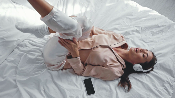 mujer feliz en auriculares inalámbricos acostado en la cama con teléfono inteligente - Metraje, vídeo