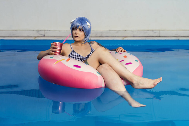 A European female with blue hair drinking a cocktail on an inflatable in a pool - Фото, изображение