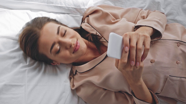 Neşeli bir kadın yatakta uzanmış akıllı telefondan konuşuyor. - Video, Çekim