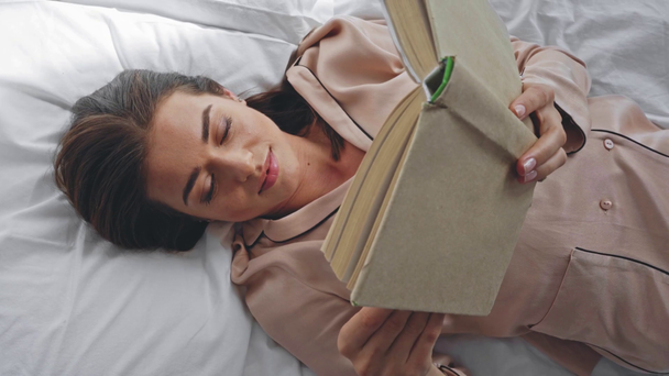 bovenaanzicht van een lachende vrouw die het boek leest terwijl ze in bed ligt - Video