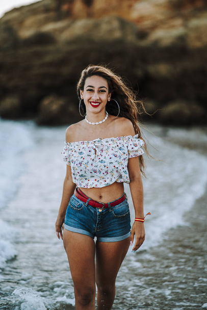 A sexy caucasian woman in short jeans and an open shirt showing a fit belly walking along a coastline - 写真・画像