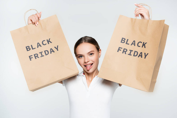 young woman sticking out tongue and holding shopping bags with black friday lettering isolated on white - Foto, Bild