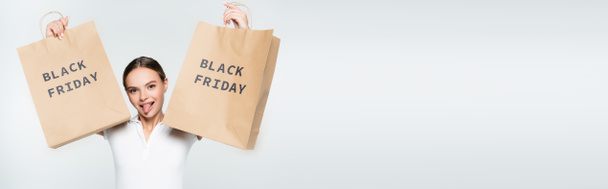 panoramic shot of woman sticking out tongue while holding shopping bags with black friday lettering on white - Фото, зображення