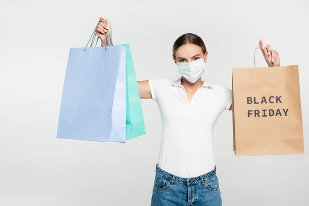 jonge vrouw in medisch masker houden boodschappentassen met zwarte vrijdag belettering geïsoleerd op wit  - Foto, afbeelding