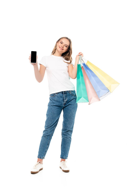 elégedett fiatal nő gazdaság okostelefon üres képernyő közelében színes bevásárló táskák elszigetelt fehér - Fotó, kép