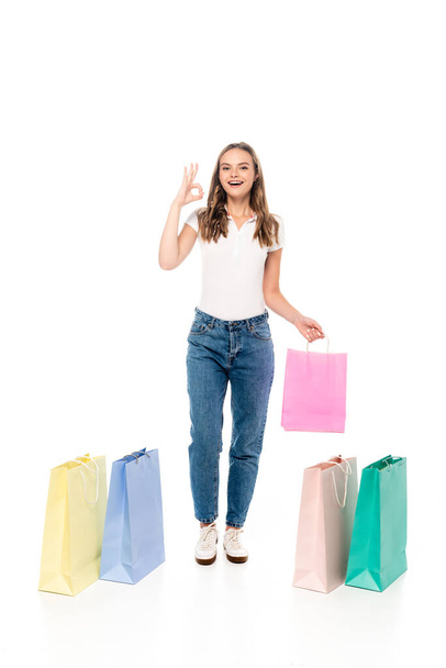 elégedett és fiatal nő mutatja rendben jel közelében színes bevásárló táskák elszigetelt fehér - Fotó, kép