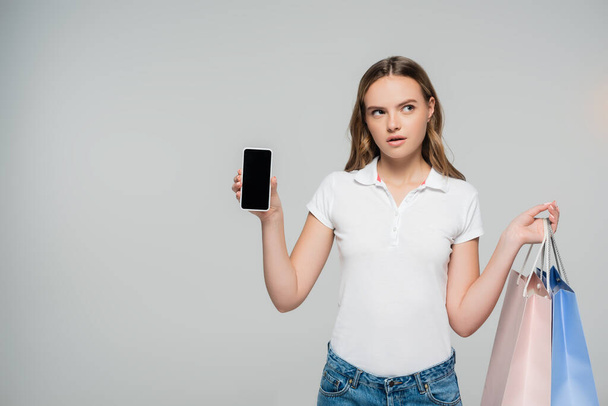 mujer pensativa sosteniendo teléfono inteligente con pantalla en blanco y bolsas de compras aisladas en gris  - Foto, Imagen
