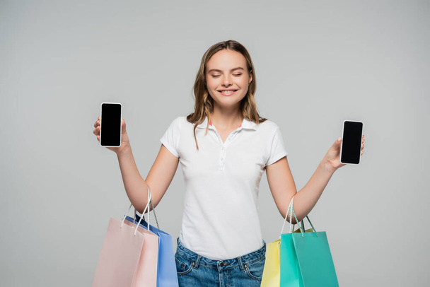 mujer alegre con los ojos cerrados sosteniendo teléfonos móviles con pantalla en blanco y bolsas de compras aisladas en gris, concepto de viernes negro  - Foto, Imagen