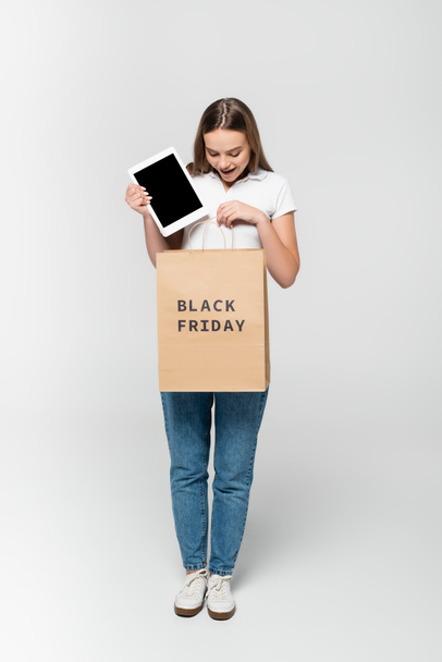 izgatott fiatal nő gazdaság digitális tabletta üres képernyőn közelében bevásárló táska fekete péntek felirat szürke - Fotó, kép