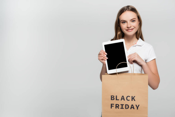 vidám fiatal nő kezében digitális tabletta üres képernyőn közelében bevásárló táska fekete pénteken felirat szürke - Fotó, kép