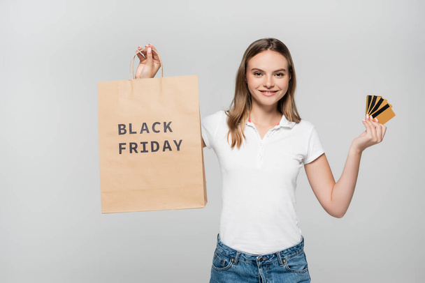 pleased woman holding credit cards and shopping bag with black friday lettering isolated on grey - Photo, Image