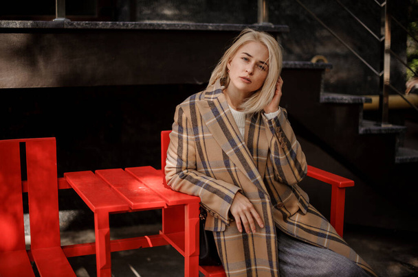 handsome young blonde woman in autumn plaid coat sitting on bright red bench - Foto, Imagem