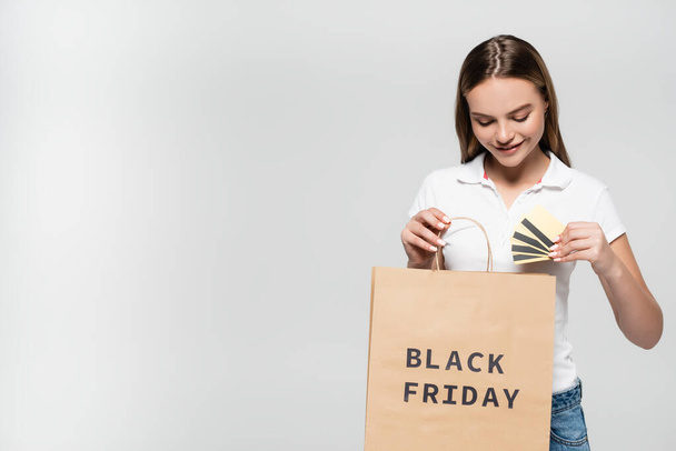 joyful woman holding credit cards and looking at shopping bag with black friday lettering isolated on grey - 写真・画像