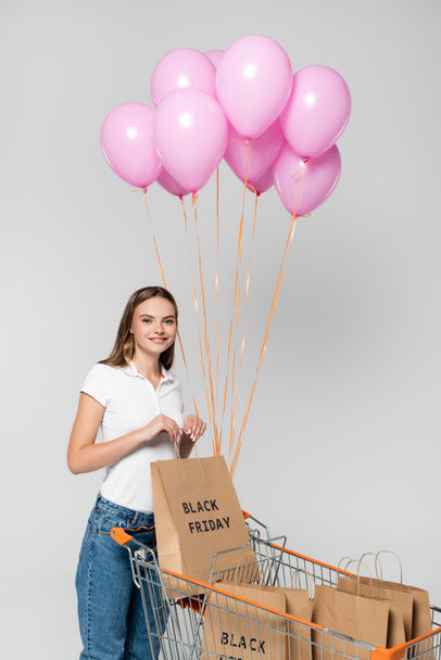 Joven mujer alegre sosteniendo bolsa de compras con letras de viernes negro en el carrito con globos rosados aislados en gris - Foto, imagen