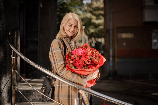 cheerful woman in plaid coat stands with bouquet of bright flowers in her hands - Фото, зображення