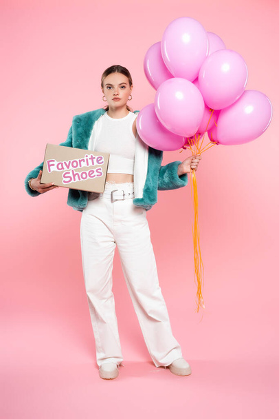 young woman holding carton box with favorite shoes lettering and balloons while standing on pink  - Foto, imagen