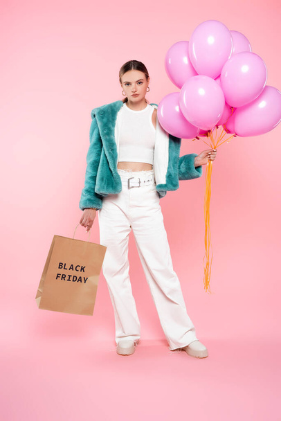 young woman holding shopping bag with black friday lettering and balloons on pink  - Foto, imagen