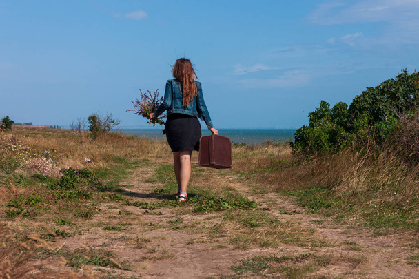 Young woman with long brown hair in denim jacket, black skirt, vintage suitcase,purple wild flowers bouquet off-road sea landscape. Hitchhiker on countryside trip. Lifestyle photo lady walking outdoor - Photo, Image