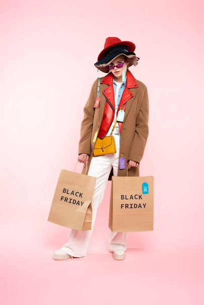 jovem mulher em óculos de sol e chapéus com etiquetas de venda de pé com sacos de compras em rosa, conceito de sexta-feira preta - Foto, Imagem