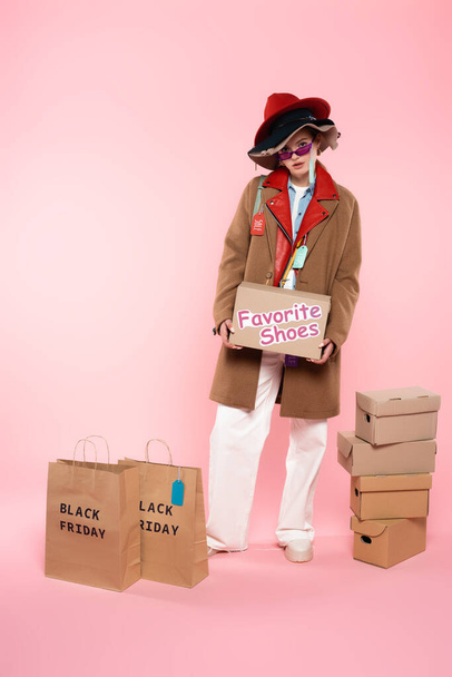 young woman in sunglasses and hats with sale tags holding box with favorite shoes lettering near shopping bags on pink, black friday concept - Photo, Image